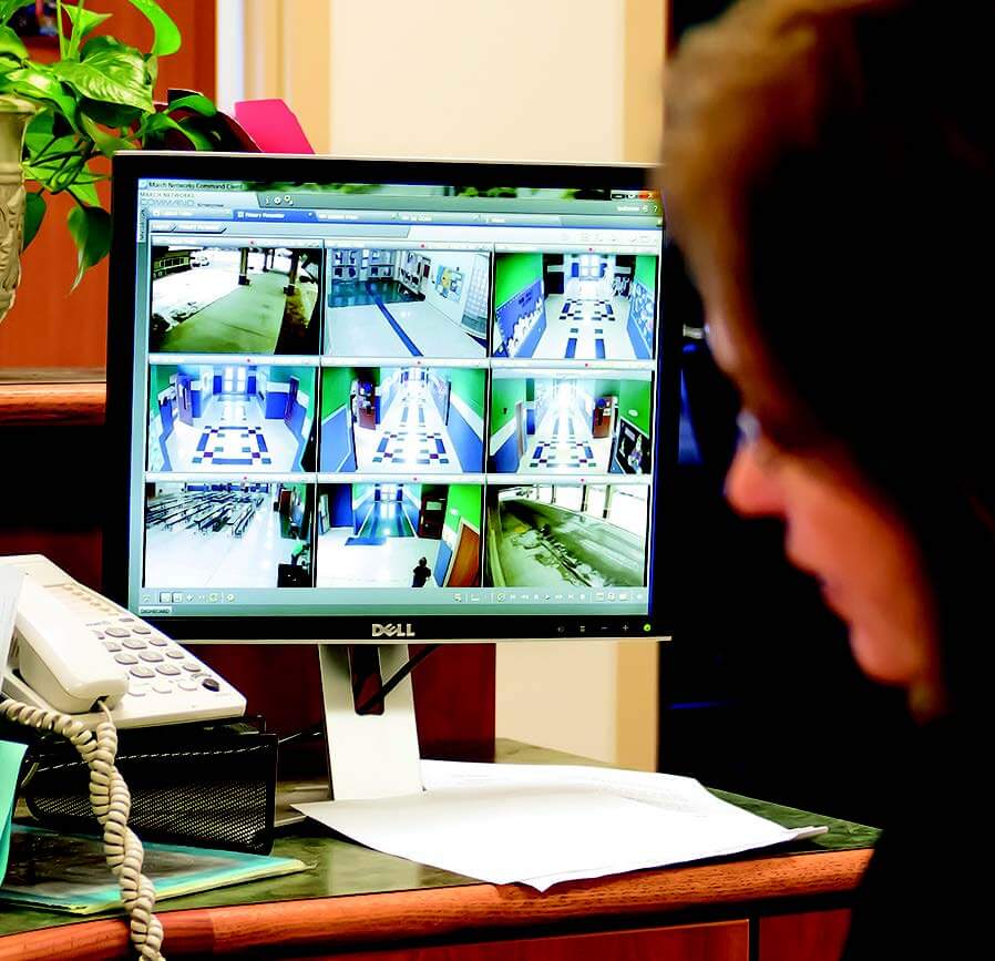 Image of a lady at a desk with security camera footage of a school on the computer screen. 