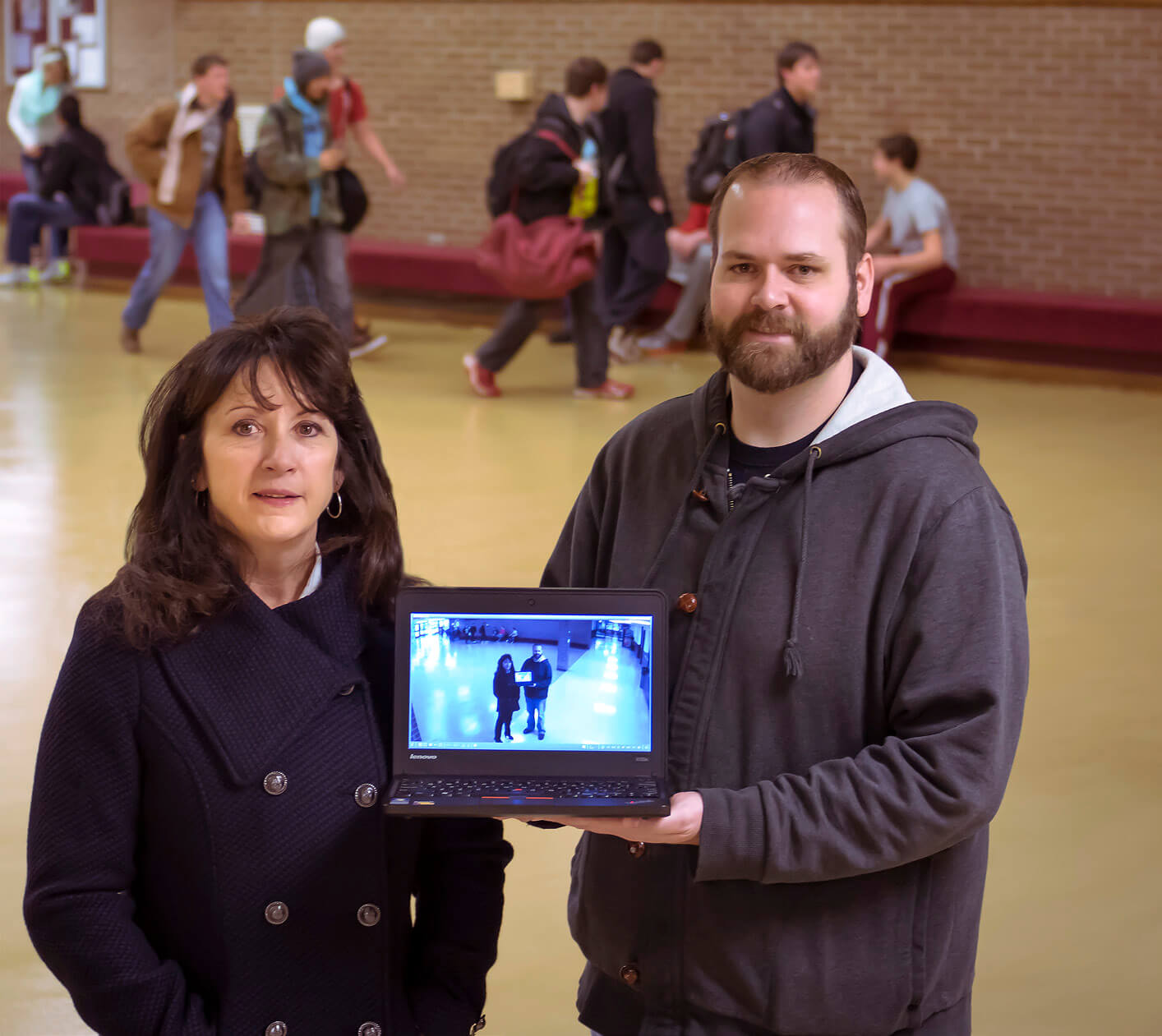 Image of two people holding up a laptop with security camera footage of themselves on the screen.