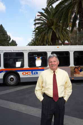 A portrait of Art Leahy, OACTA CEO, standing in front of an OCTA bus, many palm trees line a road in the background