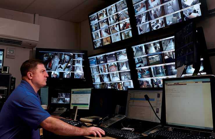 Security personnel viewing 6 large monitors with multiple views of hospital hallways, parking lots, reception etc.